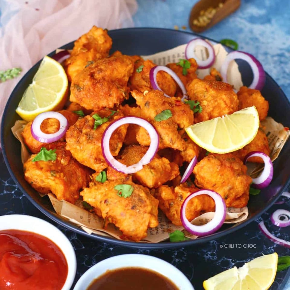 A black bowl of golden-fried Indian pakoras, garnished with red onion rings, cilantro, and lemon wedges.