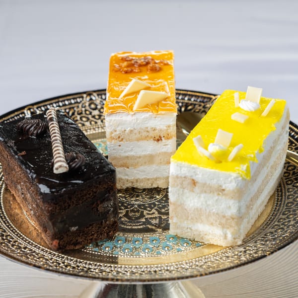 Three colorful Indian pastries, chocolate, orange, and yellow, on a decorative gold and black tray.