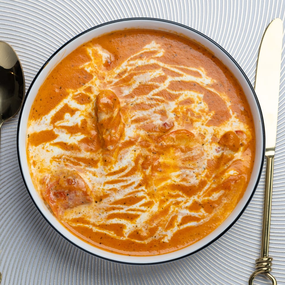 Overhead view of a bowl of Indian butter chicken, garnished with cream swirls, next to gold cutlery.