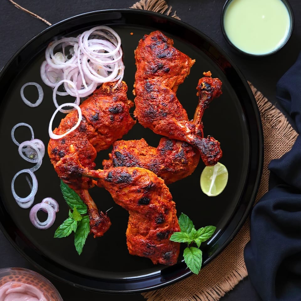 Taj Mahal Grill & Bar's Tandoori chicken, garnished with onion rings, mint, and lime, on a black plate.