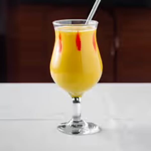 A tall glass of yellow mango lassi with red swirls and a straw on a white table.