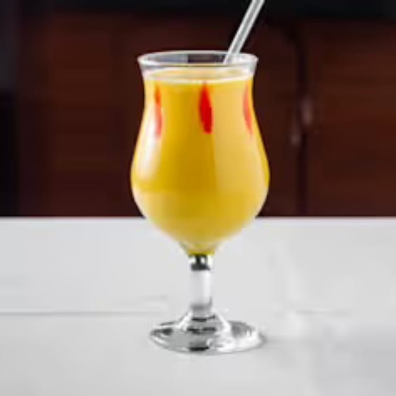 A tall glass of yellow mango lassi with red swirls and a straw on a white table.