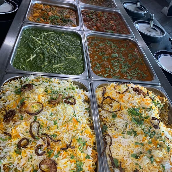 A buffet spread of Indian dishes, including biryani, palak paneer, and various curries in metal trays.