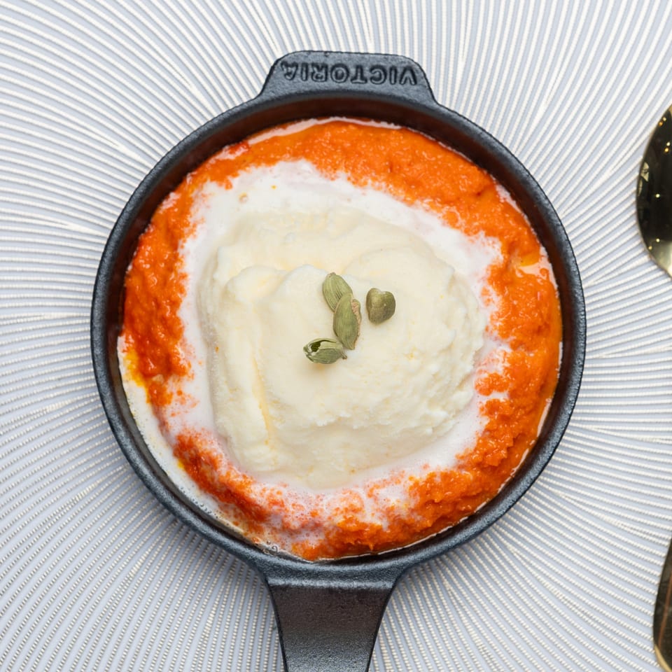 Indian dessert in a black skillet, with orange purée, white cream, and green cardamom pods.