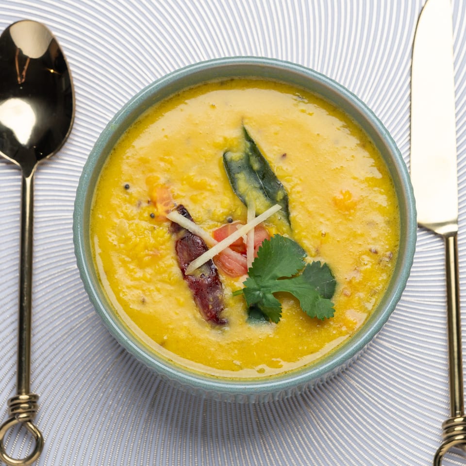 A bowl of yellow Indian dal, garnished with cilantro, ginger, and red chilies, with gold cutlery.