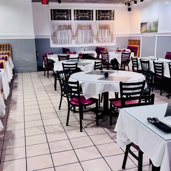 Empty restaurant dining room with white tablecloths, red chairs, and decorative stained glass windows.