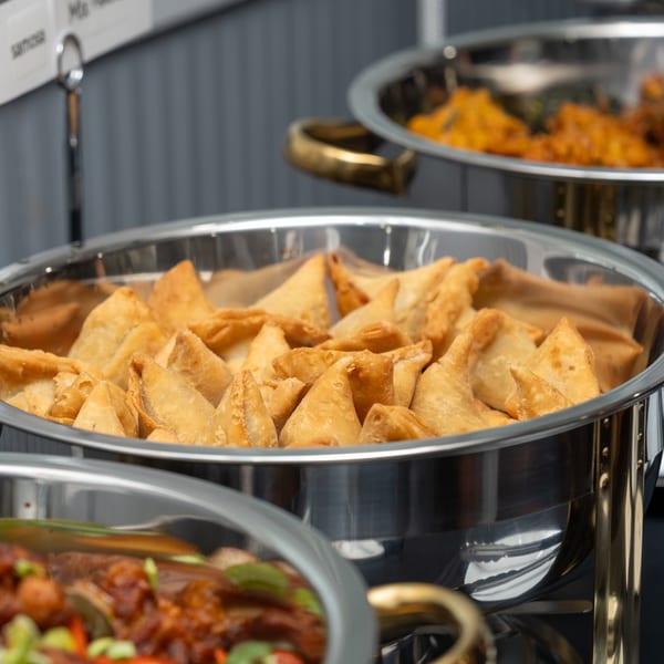 Golden-brown samosas fill a silver chafing dish, with other Indian dishes blurred in the background.