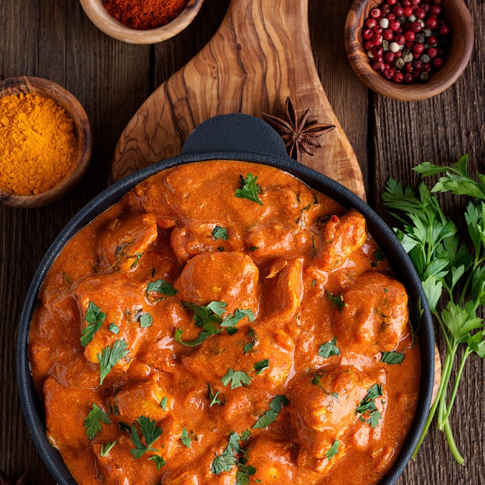 Chicken tikka masala in a black pot with spices and parsley on a wooden table.