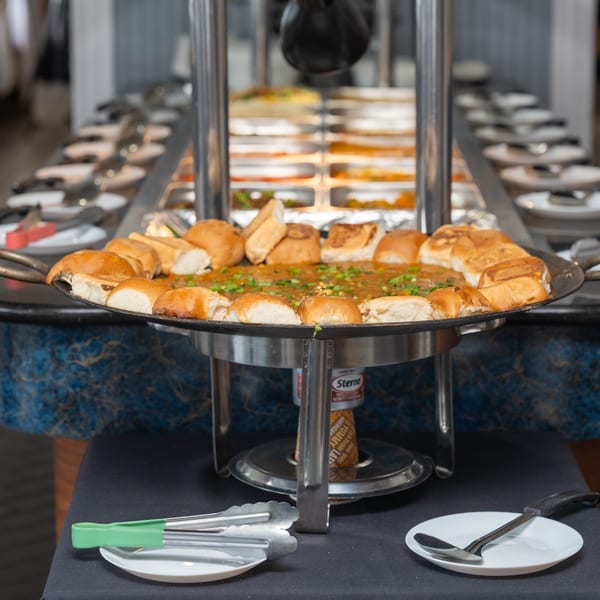 Indian buffet with a large pan of pav bhaji surrounded by bread rolls, tongs, and plates.