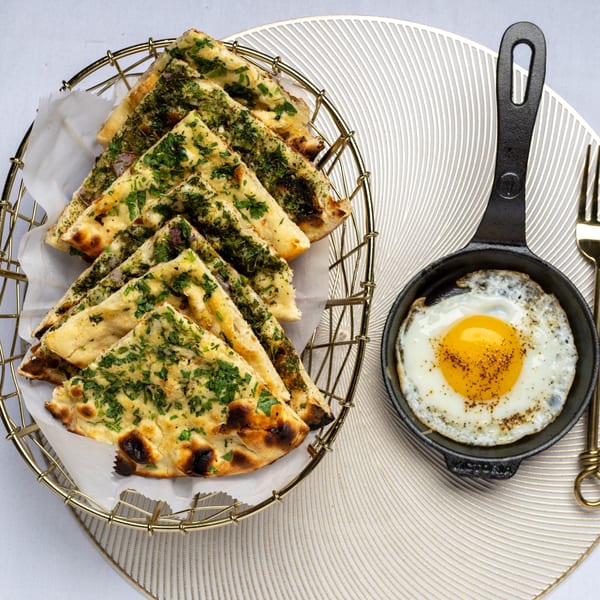 A golden basket of Indian naan bread with herbs next to a fried egg in a small skillet.