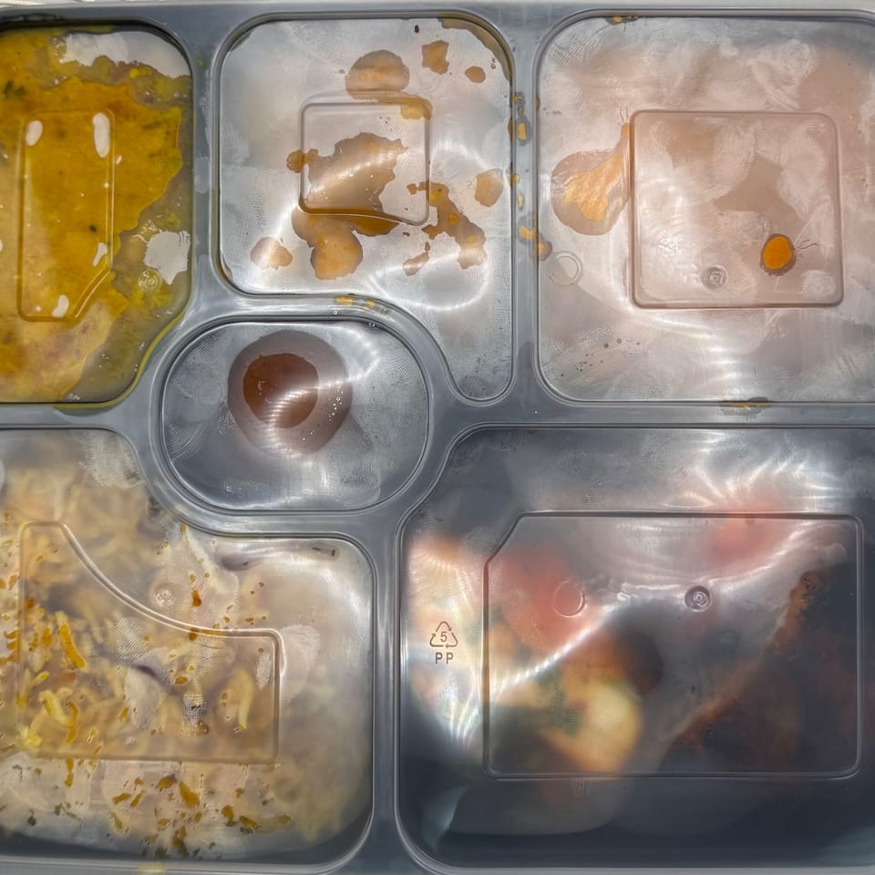 Top-down view of a plastic meal tray with five compartments of Indian food, sealed with a clear lid.