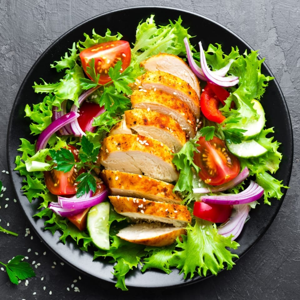 Grilled chicken breast slices with sesame seeds on a fresh green salad with tomatoes, red onion, and cucumber.