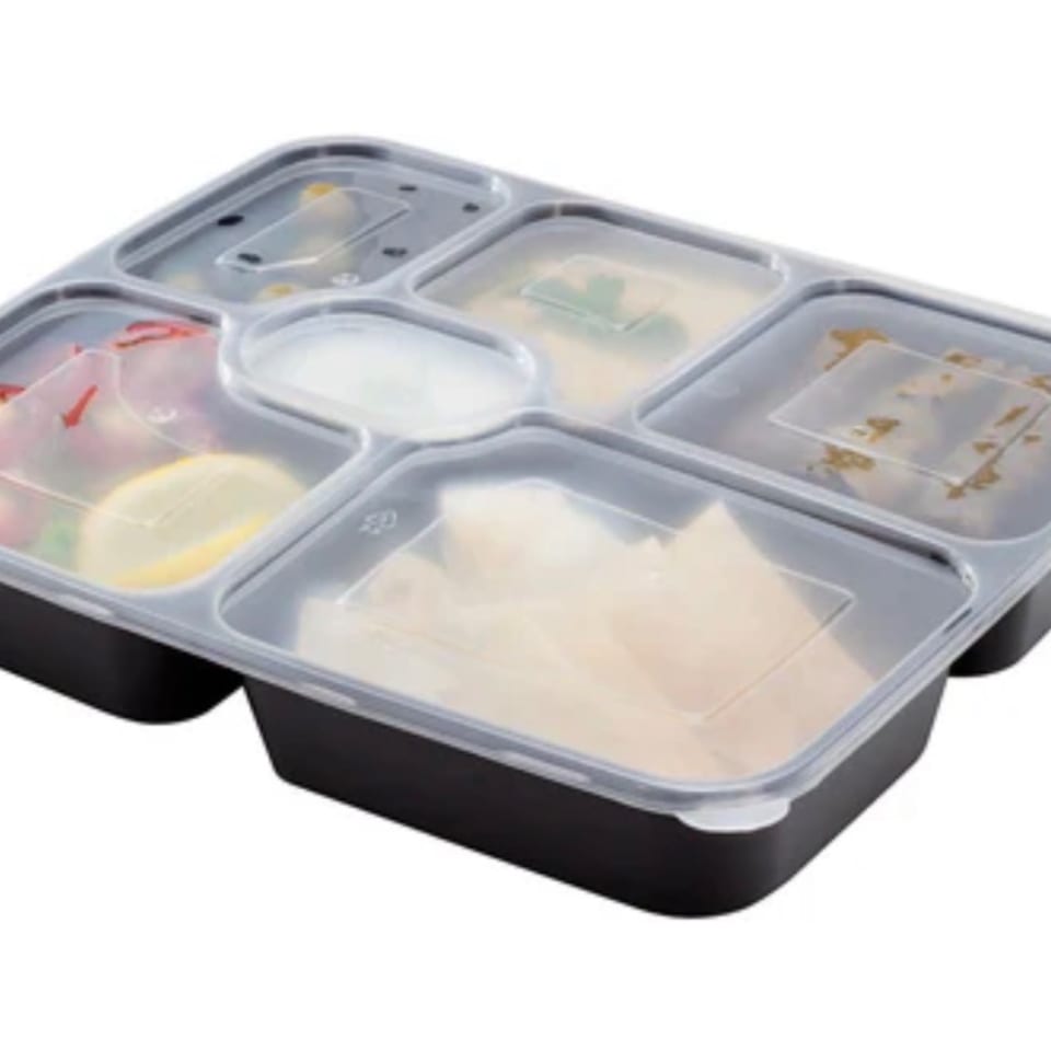 A black bento box with clear lid, filled with various food items like salad, lemon, and sauces.