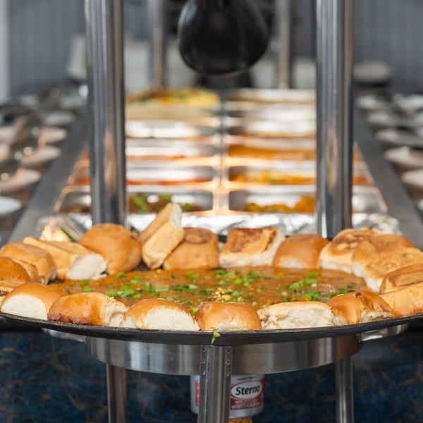 A large pan of pav bhaji, a vibrant Indian vegetable curry, surrounded by bread rolls at a buffet.