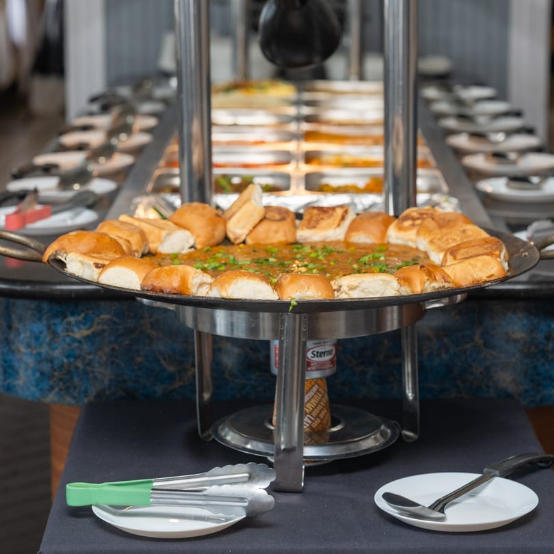 Indian buffet with a large pan of food, garnished with green onions, surrounded by bread rolls.