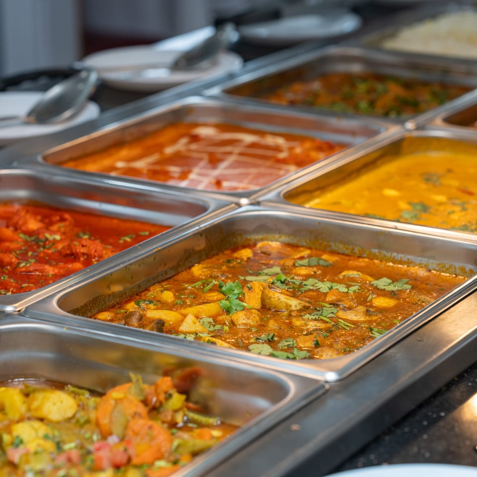 A buffet line with several metal trays filled with various Indian curries and dishes.