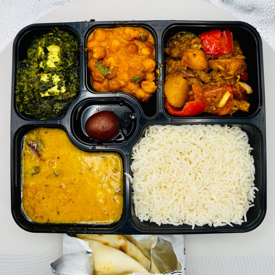 Indian thali meal in a black partitioned container with rice, dal, paneer, chana masala, and vegetables.