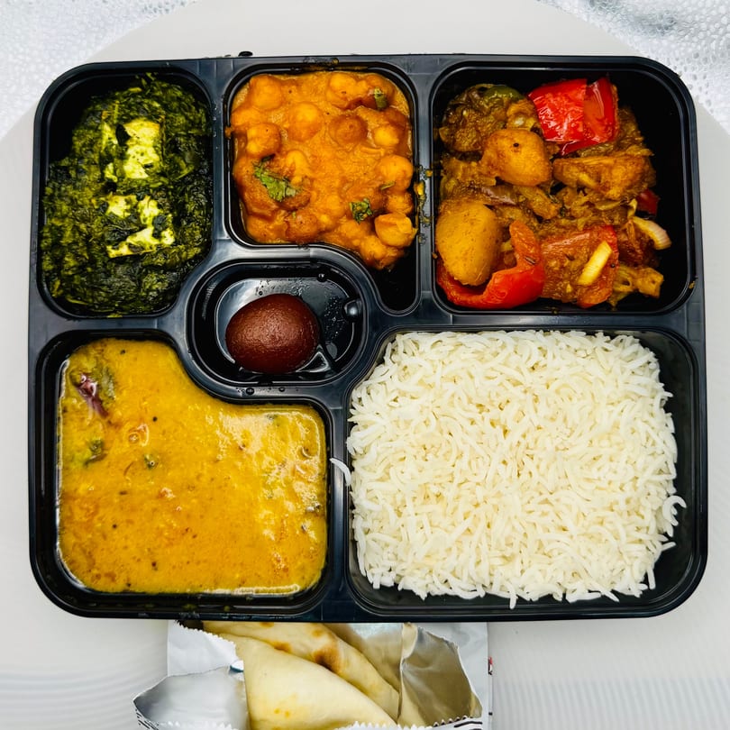 Indian thali meal in a black partitioned container with rice, dal, paneer, chana masala, and vegetables.