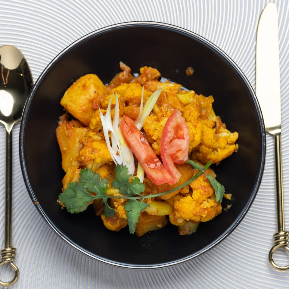 Indian cauliflower and potato curry, garnished with cilantro, tomatoes, and ginger in a black bowl.