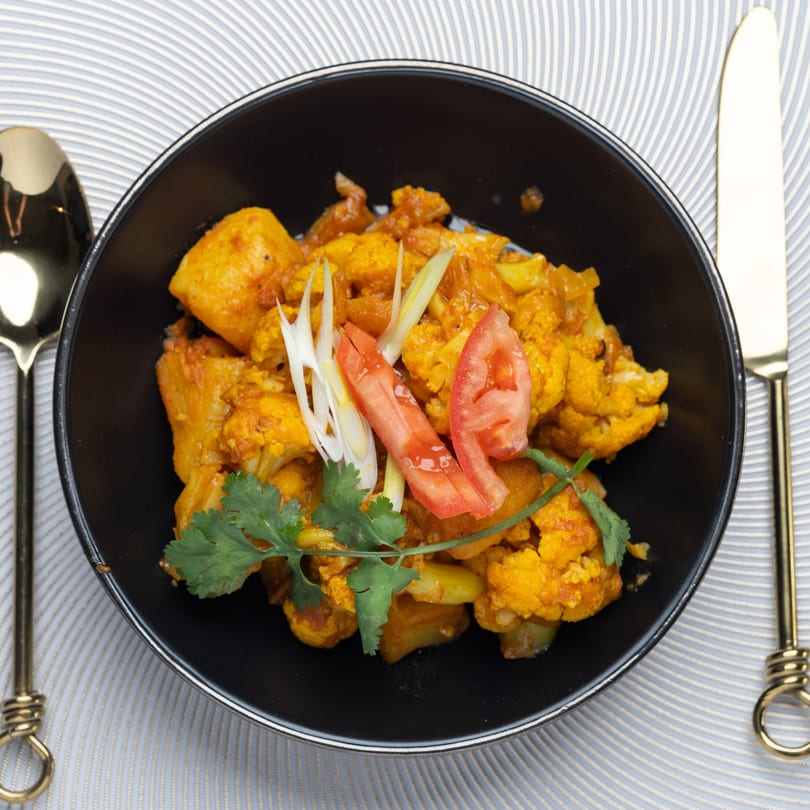 Indian cauliflower and potato curry, garnished with cilantro, tomatoes, and ginger in a black bowl.