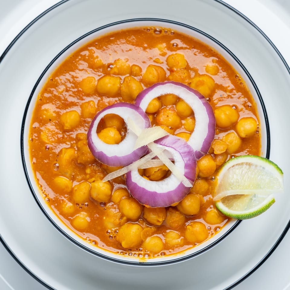 Close-up of Indian chana masala with red onion, ginger, and lime in a white bowl.