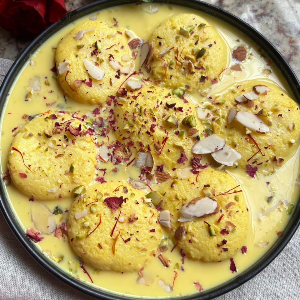 Rasmalai, an Indian dessert, with yellow patties in creamy sauce, garnished with nuts and rose petals.