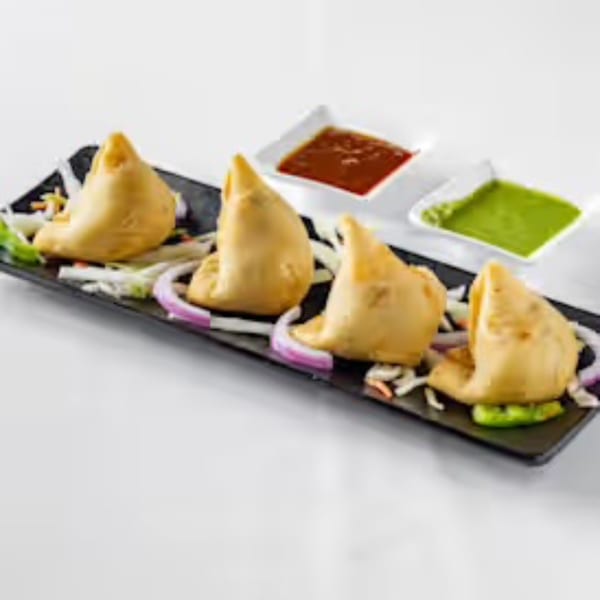 Four golden-brown samosas on a black plate with red onion, served with red and green chutneys.