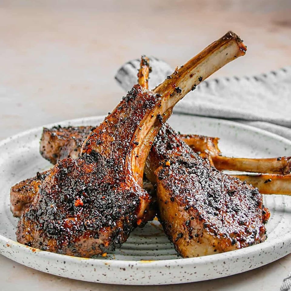 Close-up of four Indian-spiced lamb chops with visible bones, coated in dark seasoning on a white plate.